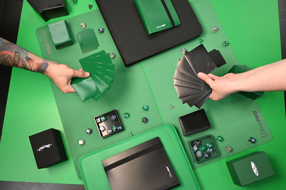 An overhead view of a gaming setup featuring a green and black Magic: The Gathering playmat, cards, dice, and various game accessories arranged on a bright green surface. Two hands are visible holding fanned decks of cards.